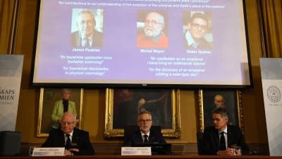 Tre män sitter framför ett skrivbord och berättar vem som vunnit Nobelpriset i fysik. Bakom dem syns bilderna på de tre vinnarna.