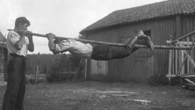 En man håller i en stång med två händer och sina fötter och hänger i den. Bilden är tagen 1930 i Sund, Åland.
