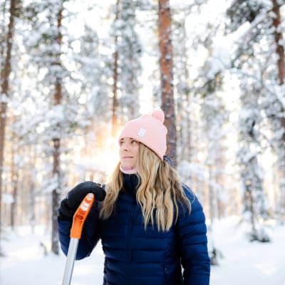 Nainen seisoo lumisessa metsässä nojaten lumilapioon.