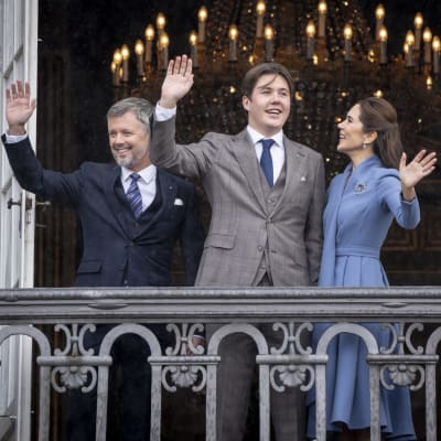 Kronprins Frederik, prins Christian och kronprinsessan Mary ler och vinkar stående på en balkong.