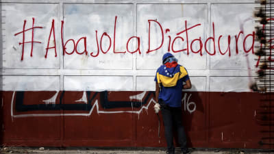 En demonstrant håller i stenar i sammandrabbning med polis i Venezuela. På väggen står "Ner med diktaturen" på spanska.  