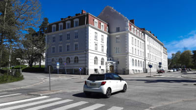 En vit byggnad bakom en korsning. Det är en solig dag. En bil kör genom korsningen.