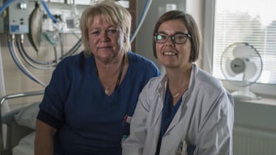 Två kvinnor inom sjukvården sitter på en sjukhussäng.