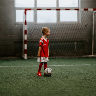 Ett barn står med en fotboll framför sig på en fotbollsplan inomhus.