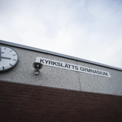 En skolvägg med en klocka, en megafon och en skylt. På skylten står det "Kyrkslätts gymnasium"