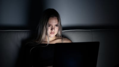 Ung flicka med långt hår sitter i ett mörkt rum, ansiktet lyses upp av dataskärm