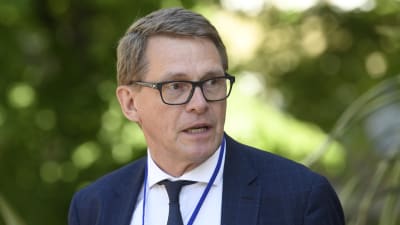 Finansminister Matti Vanhanen blickar snett i kameran med öppen mun. Han står framför gröna buskar och har slips och kostym på sig.