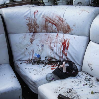 Blodfläckar på en vit soffa av läder. På soffan finns också en svart keps som är blodig och en tom vattenflaska.