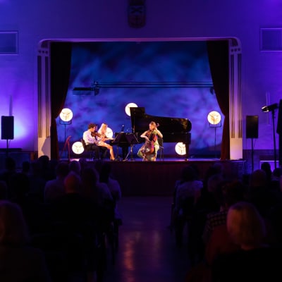 Tre musiker uppträder framför publik. Salen är mörklagd och scenen belyst i ett lila sken. Musikerna spelar violin, piano och cello.