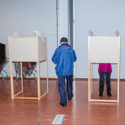 Kaksi äänestyskoppia huoneessa, toisessa kopissa on äänestäjä, joka on pukeutunut punaiseen takkiin, toiseen äänestyskoppiin on matkalla henkilö jolla on sininen takki.