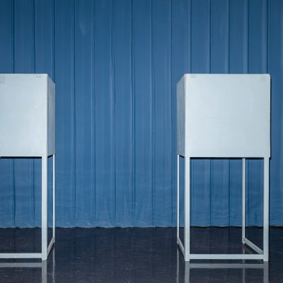 Kaksi sinisenharmaata äänestyskoppia huoneessa, takaseinällä on sininen koko seinän peittävä verho.