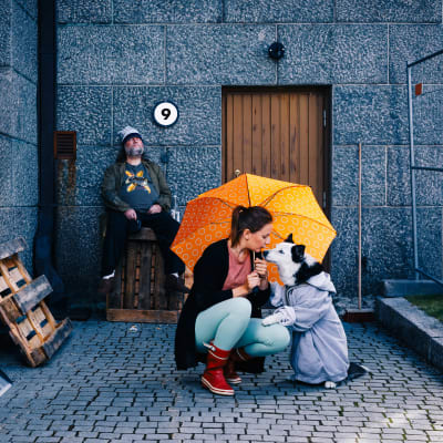 Nainen ja koira ovat oranssisen sateenvarjon alla. Partainen mies istuu taustalla seinän vieressä.