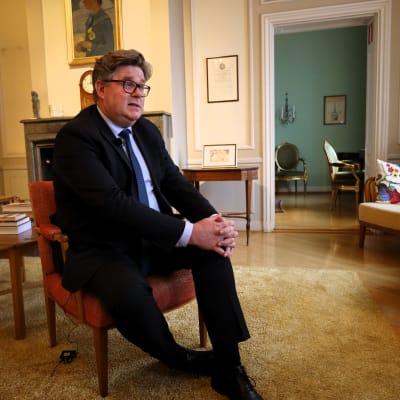 En man i mörkkostym sitter på en stol i elegant salong på svenska ambassaden i Helsingfors. 