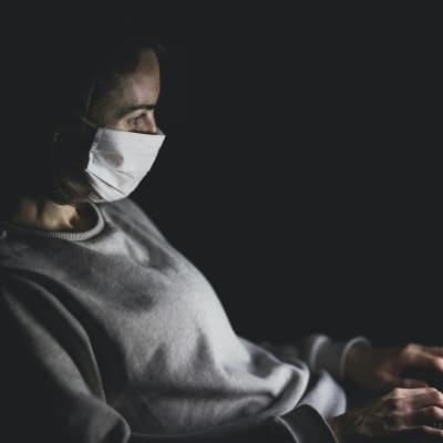 Kvinna med munskydd över näsa och mun sitter i mörkret och skriver på bärbar dator.