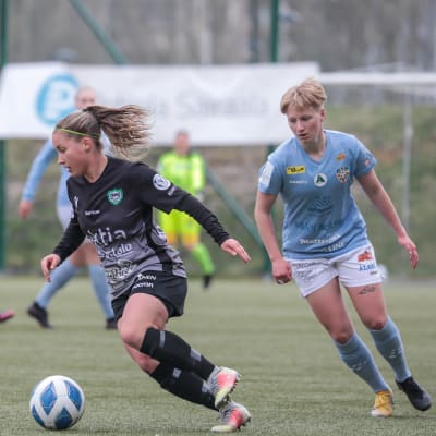 Två kvinnliga fotbollsspelare löper på planen. Spelaren klädd i svart har bollen, spelaren i blått följer efter.