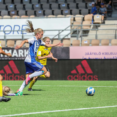Linda Sällström i blåvitrandig tröja driver med bollen mot KuPS. Hon jagas av KuPS-spelare i gula tröjor. En HJK-spelare har fallit till marken.