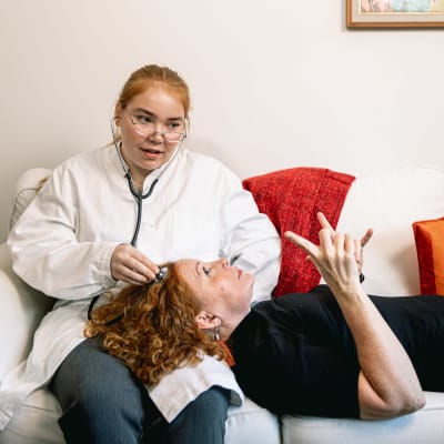 Heidi ligger i soffan med huvudet i Vivis knä. Vivi håller ett medicinskt stetoskop mot Heidis tinning som under en undersökning.