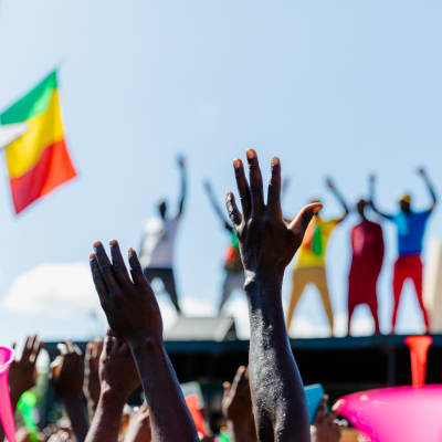 Kaksi kättä ilmassa, taustalla jonkinlaisen katoksen päällä seisovien ihmisten hahmot, rakennuksen torni ja Malin lippuja. 