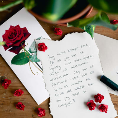 Ett kärleksbrev skrivet för hand intill ett kuvert adresserat till "älskling" på ett bord med rosor.