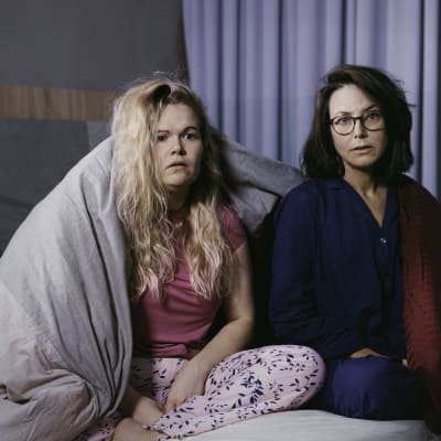 Två kvinnor sitter på en säng iklädda pyjamas och ser förskräckta ut. Ett täcke hänger över ena axeln på kvinnan till vänster.