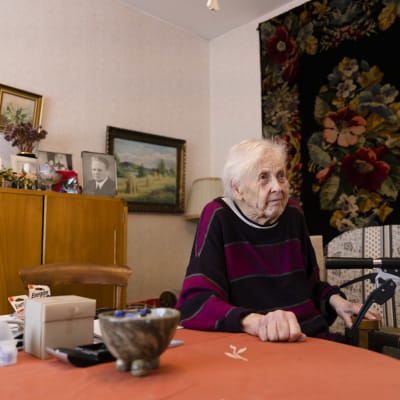 Vanha nainen kotonaan. Seinillä on ryijy ja tauluja, nainen istuu pöydän äärellä. 