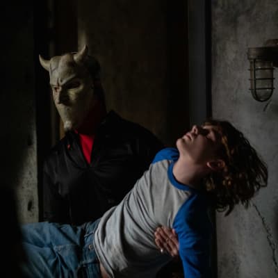 En maskerad figur bär en medvetslös pojke i ett skumt rum. 