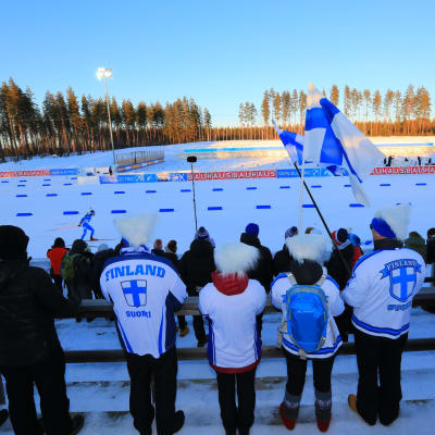 Yleisö heiluttaa siniristilippuja ampumahiihtostadionin katsomossa.