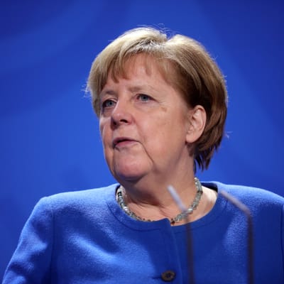 Angela Merkel, blå tröja framför en blå bakgrund. Framför mikrofoner.