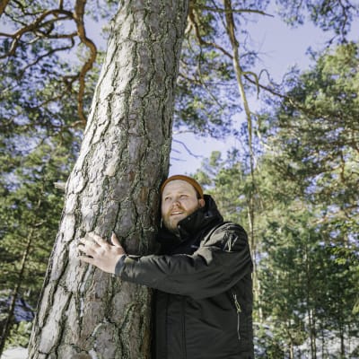 Jonas Forsbacka står och kramar ett träd ute i skogen på Emsalö. Jonas har en ängslig min, och skogen syns i bakgrunden. 