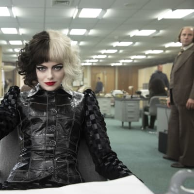 Närbild på Cruella (Emma Stone) som sitter och ser klurisk ut.