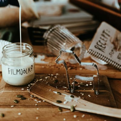 Ett smutsigt köksbord med en öppen receptbok, en pepparkaksform och en glasburk som fylls av en vit vätska. På burken står det "filmjölk" med frågetecken.