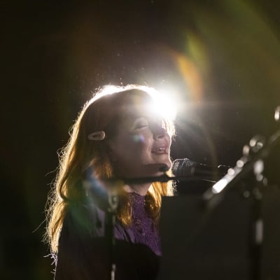 Astrid Swans ansikte, orange hår, en strålkastare som lyser bakom henne. Hon sjunger i en mikrofon med slutna ögon.