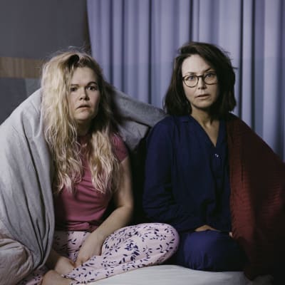 Två kvinnor sitter på en säng iklädda pyjamas och ser förskräckta ut. Ett täcke hänger över ena axeln på kvinnan till vänster.