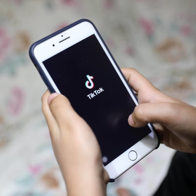 En person håller i en vit telefon. I skärmen syns Tiktoks logo; svart bakgrund med en not och texten "Tiktok".