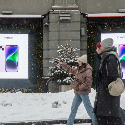 Ihmisiä kavelee lumisella kadulla. Taustalla iphone-puhelimen mainos.