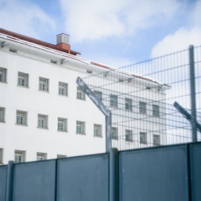 En ljus fängelsebyggnad med ett staket i förgrunden och rader av gallerförsedda fönster på andra sidan staketet.