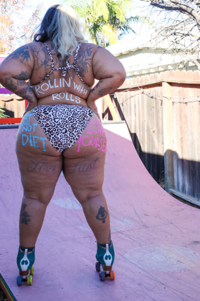 En kvinna med rullskridskor står med ryggen till. Hon är iklädd bikini, och på kroppen står slagord målade i olika färger: Riot not diet, Rollin' with rolls och Love yourself 