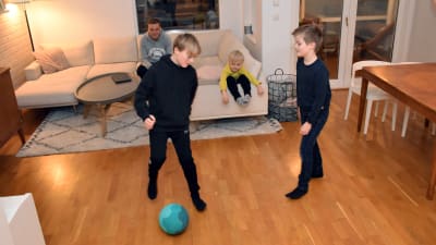 Två pojkar spelar fotboll i ett vardagsrum, pappan och yngsta pojken sitter i soffan. 