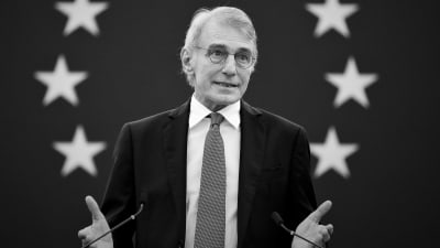 En svartvit bild av David Sassoli som pratar. Han står framför en vägg som har EU:s flagga på sig.