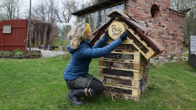 Blond kvinna hänger upp skylt med texten Bug Inn på ett litet hus byggt av lastpallar och taktegel.