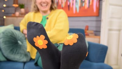  Nainen istuu sohvalla jalat ojennettuna pöydällä. Hänen sukkansa on paikattu värikkäillä kukilla.
