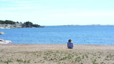 Kvinna med ryggen mot kameran sitter på en sandstrand och ser ut över havet en varm sommardag.