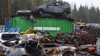 Skrotbilar på en industrigård. Där finns också annat metallskrot. I mitten en stor grön container som det står Romu Keinänen på.