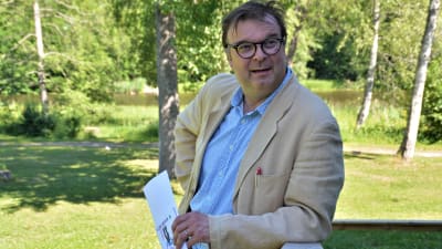 Man i skjorta och kavaj lutar mot en ledstång på en trappa utomhus. I handen har han en broschyr. I bakgrunden gräsmatta, träd och en sjö.