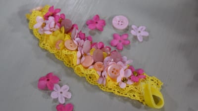 Ett armband av gul spets med rosa detaljer som små tygblommor, udda knappar och pärlor.