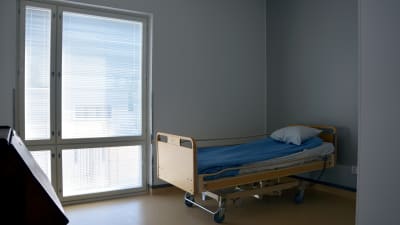 En säng för en äldre person står nära ett fönster i ett rum.