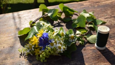 En halvfärdig blomsterkrans där små buketter av blåklint, björkris, prästkrage, allium och daggkåpa bundits på en björkkrans med hjälp av en tunn metalltråd.
