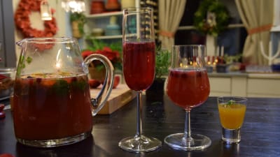 En glaskanna med röd dryck med fruktskivor och bär i, samt ett glas champagne med kall glöggdrink, ett vinglas med tranbärsdryck och ett snapsglas med gul äppelshot.