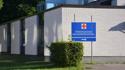 Hangö hälsocentral, en vit tegelbyggnad i 1-2 våningar. Soligt väder, grönt gräs. En blå skylt där det står Terveyskeskus Hälsovårdscentral.