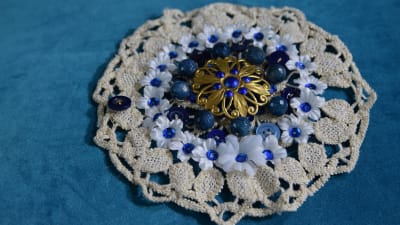 Ett stort armband av en återvunnen virkad duk med vita tygblommor, blåa paljetter, knappar och pärlor.